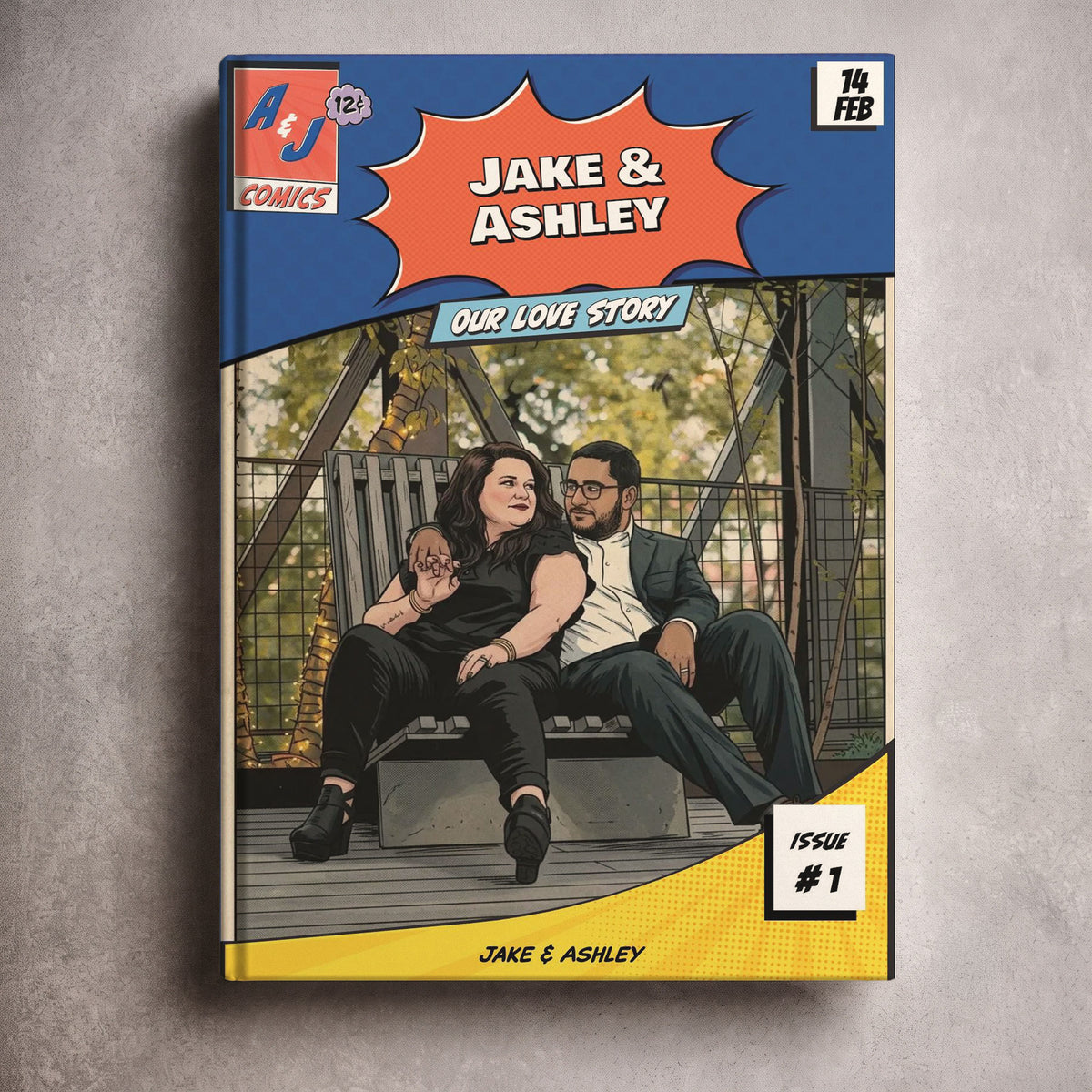 A physical comic book titled 'Jake & Ashley: Our Love Story' from 'AJ Comics' is shown standing upright on a textured gray surface. The cover features a blue top section with the title, an illustration of a couple sitting on a bench, and a yellow bottom section with the issue number.